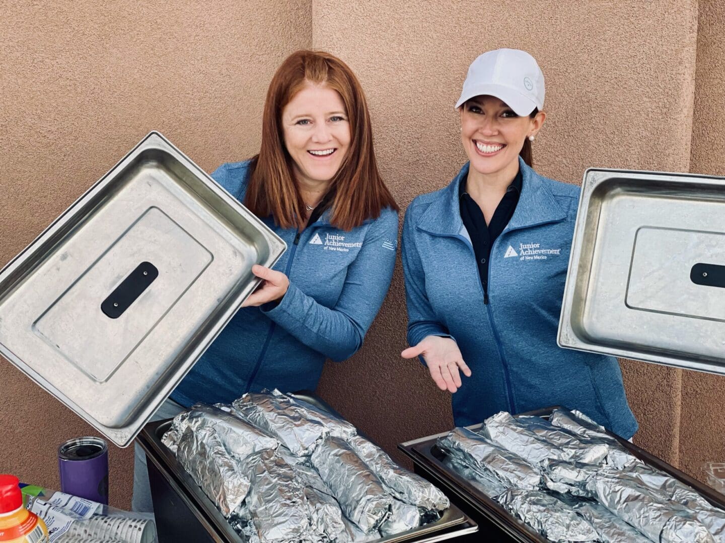 Two women holding trays of food in front of a wall.